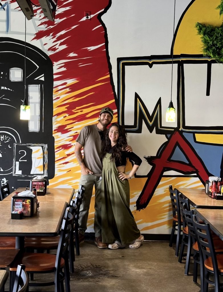 Two guests posing in front of the Mullets Ames mural inside the restaurant