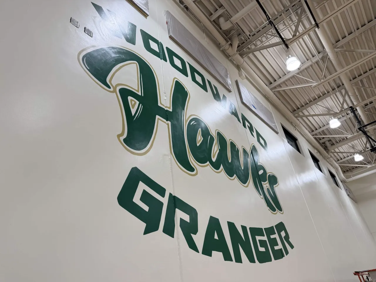 The Woodward Granger Hawks mural seen from the left side of the gym, with white ceiling trusses above it