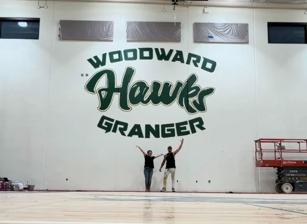 Two people standing beneath the finished Woodward Granger Hawks mural with their arms raised